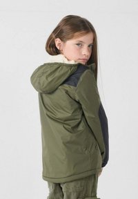 Olijfgroene jas met capuchon en zwarte accenten, gevoerd met nepbont. Soepele textuur, elastische manchetten en een iets losse pasvorm.