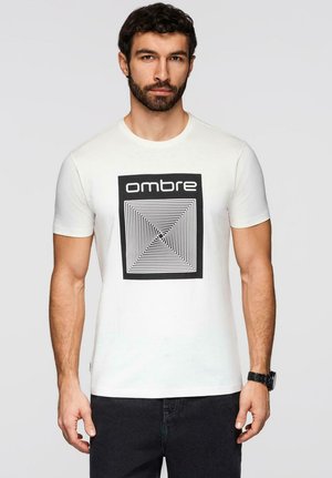 Mann mit dunklem, lockigem Haar und Bart, der ein weißes T-Shirt mit schwarzem grafischem Quadrat und dem Aufdruck "ombre" trägt, dazu dunkle Hose und eine schwarze Armbanduhr.