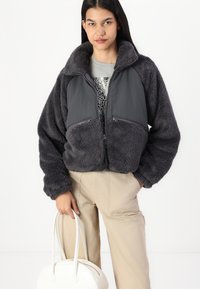 Giacca grigia, sfocata e corta con colletto alto e due tasche frontali con zip. Abbinata a pantaloni color crema e una borsa bianca.