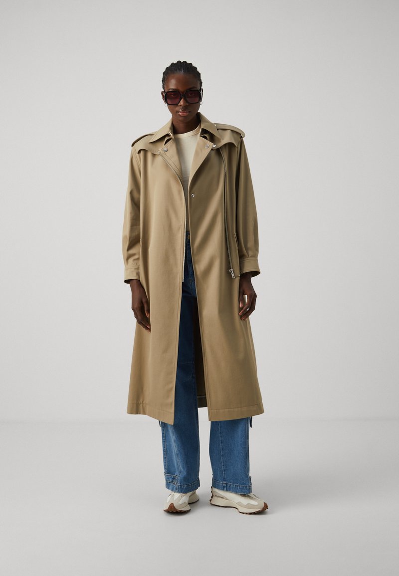 Beige Trenchcoat mit großem Kragen, Frontreißverschluss und seitlichen Taschen, über einem cremefarbenen Oberteil und blauen Jeans getragen, kombiniert mit klobigen Turnschuhen.