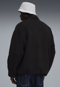 Sort fleecejakke med høj krave og oversize pasform, tekstureret overflade, parret med en hvid bucket hat, denimjeans synlige nedenfor.