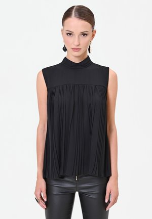 Donna con top nero plissettato senza maniche con collo alto e pantaloni in pelle neri, in piedi davanti a uno sfondo chiaro e semplice.