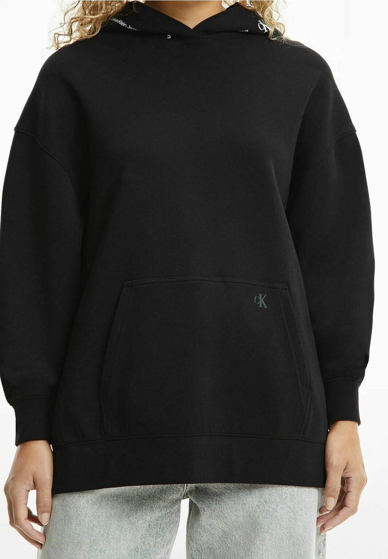 Sweat à capuche noir en tissu doux avec une poche avant, coupe ample et un logo subtil. Manches longues et poignets côtelés.