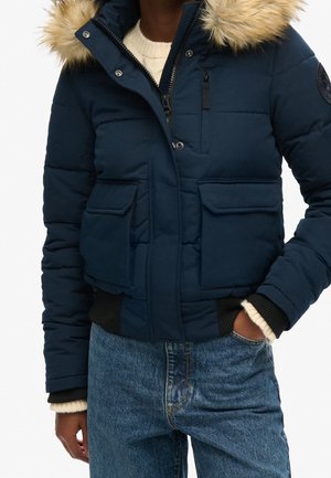 Doudoune bleu marine avec capuche bordée de fourrure, fermeture éclair et boutons-pression, poches avant à rabat, portée avec un pull léger et un jean bleu.