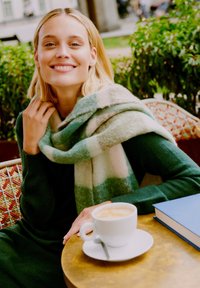 eșarfă din lână cu carouri în nuanțe de verde și bej, așezată peste un pulover de culoare verde închis. Prim-plan al unui pahar cu cafea pe o masă rotundă de aur, cu o carte aproape.