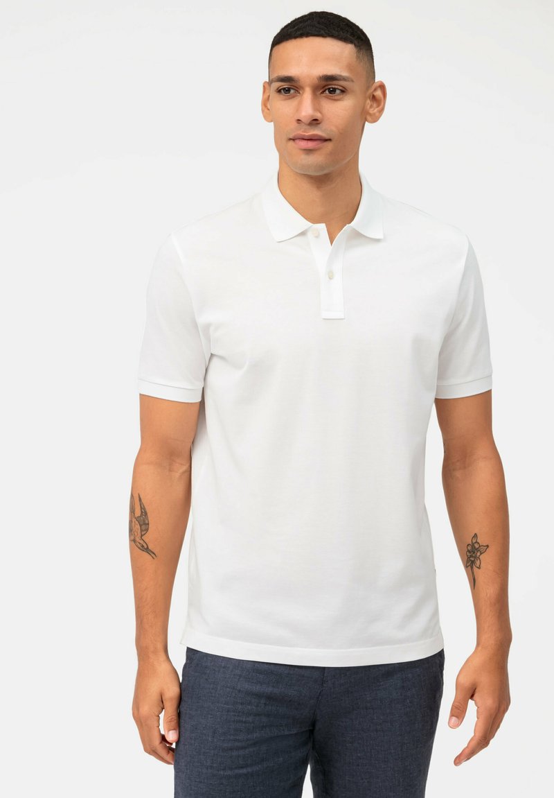Weißes Kurzarm-Polohemd mit einem Kragen-Design und einer geknöpften Halsleiste. Das Model trägt navyfarbene strukturierte Hose und hat ein Tattoo am Arm.
