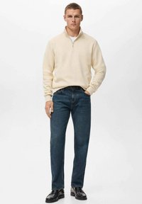 Beige zip-up sweater, blue jeans, black shoes. The sweater features a high collar and ribbed cuffs; jeans have a straight fit and slightly faded texture.