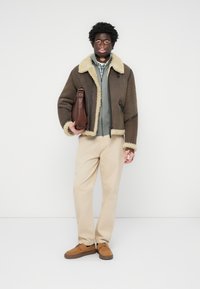 Chaqueta de borreguito marrón forrada, suéter gris con cremallera, pantalones beige y zapatos de ante marrones. Modelo sosteniendo un bolso marrón, de pie contra un fondo blanco.