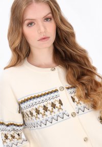 Cardigan crem cu un design modelat, care prezintă accente galbene, maro și albastre. Capsele aurii și manșetele riburate completează aspectul.