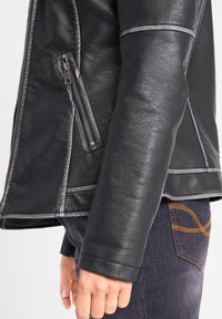 Svart läderjacka med en dragkedjeficka, kontrasterande sömmar och strukturerad design; bärs över mörka jeans med synliga gulddetaljer.