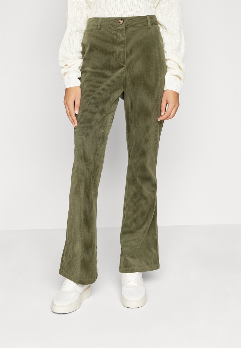 Personne portant un pantalon évasé en velours côtelé vert olive, un pull en tricot blanc et des baskets blanches, debout devant un fond clair uni.