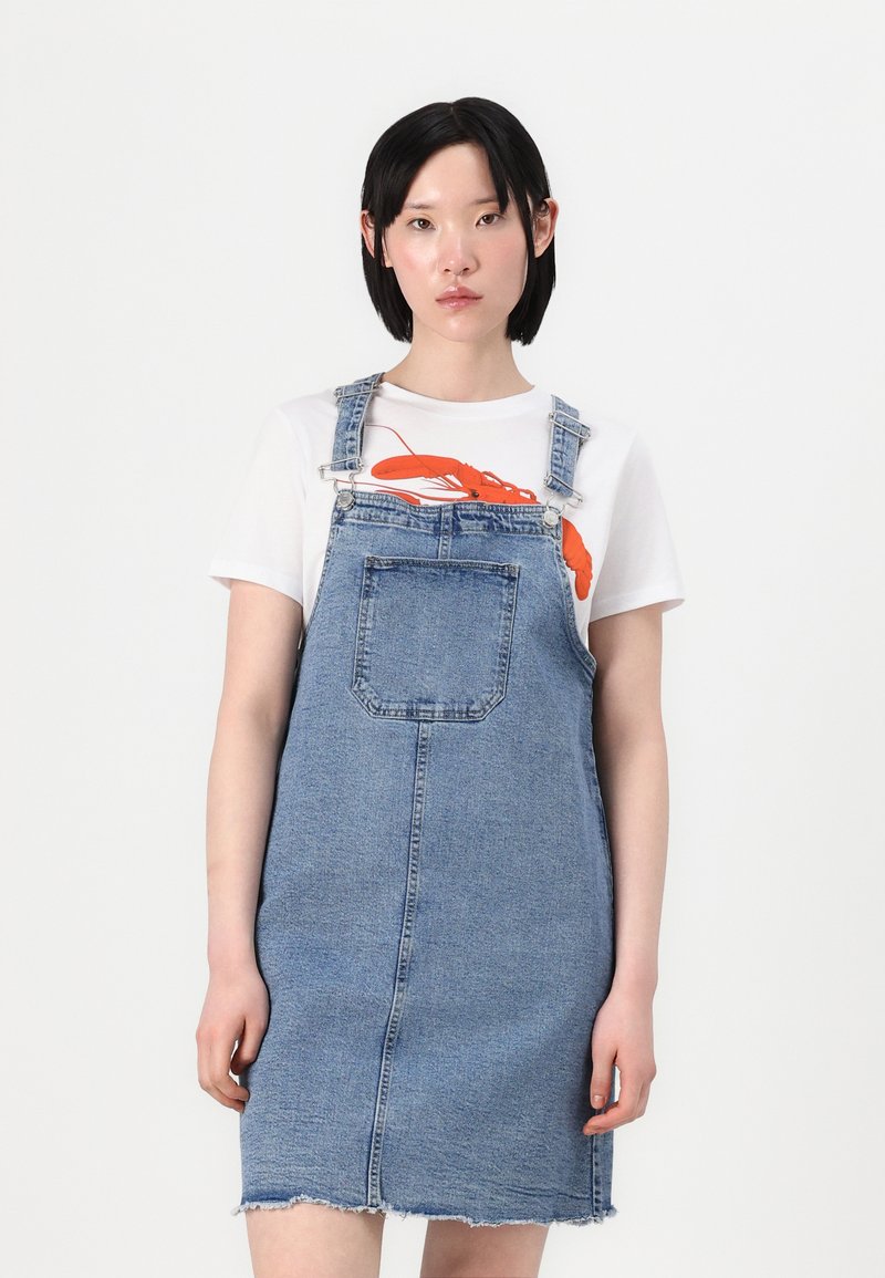 Denim kopējā kleita, gaiši zila, vidusgriezuma garumā, priekšpusē kabata, saplēsts apakšmalas dizains, uzvilkta virs balta T-kreklu ar oranžu omāra grafiku.
