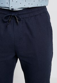 Närbild av marinblå linnebyxor med elastisk midja och snörning, i kombination med en ljusblå skjorta med små triangelmönster.