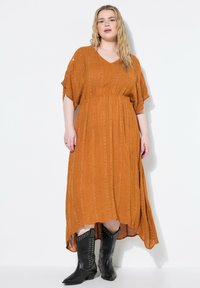 Maxikleid in rostorange mit V-Ausschnitt, kurzen Ärmeln und strukturierten Streifen. Kombiniert mit schwarzen, spitzen Cowboy-Stiefeln.