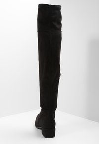 Botte en suède noire à hauteur de genou avec un petit talon carré, fermeture éclair latérale visible, photographiée de dos sur une surface blanche.