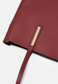 Roh burgundské texturované kožené tašky se štíhlým popruhem a zlatým obdélníkovým kovovým akcentem na bílém pozadí.