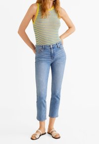 Mujer con pantalones vaqueros cortos azules, top de punto sin mangas con ribete amarillo y sandalias beige, de pie con las manos en las caderas.
