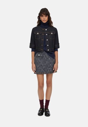 Donna con giacca corta in denim scuro sopra dolcevita blu scuro, minigonna in tweed con dettagli a bottoni, calzini bordeaux e mocassini neri.