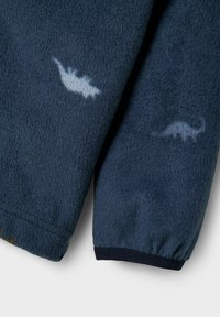 Veste en polaire bleu marine avec des motifs de dinosaures bleu clair. Dotée de poignets élastiques et d'une surface douce et texturée.