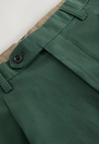 Gros plan sur un pantalon texturé vert foncé avec un rabat boutonné et une ceinture intérieure beige visible.