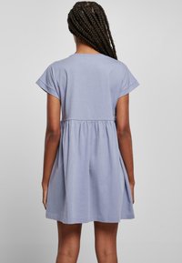 Person mit langen geflochtenen Haaren, die ein kurzärmeliges, lockeres lila Kleid trägt, von hinten vor einem schlichten grauen Hintergrund gezeigt.