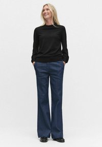 Mustat pitkät hihat -neule, jossa on korkea kaulus, yhdistettynä laivastonsinisiin leveälahkeisiin housuihin. Malli seisoo kädet taskuissa, jalassa mustat kengät.