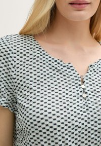 Top à manches courtes en menthe clair avec un motif en grille noir. Présente un col en V et trois boutons au centre. Tissu doux et texturé.