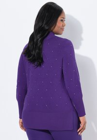 Žena s dlouhými černými vlasy, oblečená v fialovém svetru se vzorem diamantů a drobnými korálkovými detaily, zobrazená zezadu na bílém pozadí.