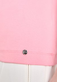 Roze katoenen t-shirt met een ronde, zilveren logo label nabij de zoom. De stof heeft een zachte textuur en een geribbelde zoomafwerking.