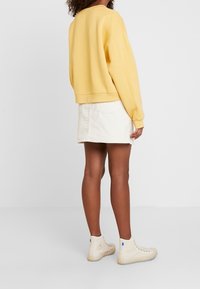 Felpa gialla corta con maniche lunghe, abbinata a una gonna in velluto a coste bianca e a sneakers beige alte. Sfondo neutro.