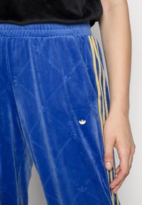 Blå sammetsbyxor med diagonalt ribbat mönster, gula sidosträck och en guldbroderad logotyp på vänster ben. Elastisk midja synlig.