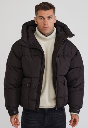 Homme avec des tatouages portant une veste matelassée noire par-dessus un pull à col roulé blanc et un jean foncé, debout devant un fond gris uni.