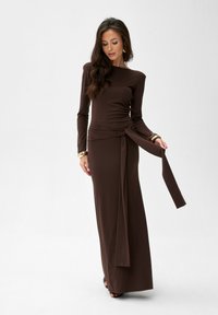 Brązowa sukienka z długim rękawem, dopasowaną górą i wiązanym detalem w talii. Gładki materiał, długość do podłogi i minimalistyczny design.