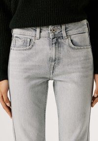 Ανοιχτό γκρι τζιν με στενή εφαρμογή, διαθέτοντας κλείσιμο με κουμπί, μπροστινές τσέπες και απαλές ξεβαμμένες λεπτομέρειες κατά μήκος της υφής του υφάσματος.