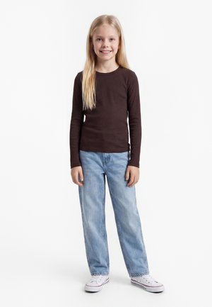 Smilende ung pige med langt blond hår iført en mørkebrun langærmet skjorte, lyseblå jeans og hvide sneakers, stående mod en hvid baggrund.