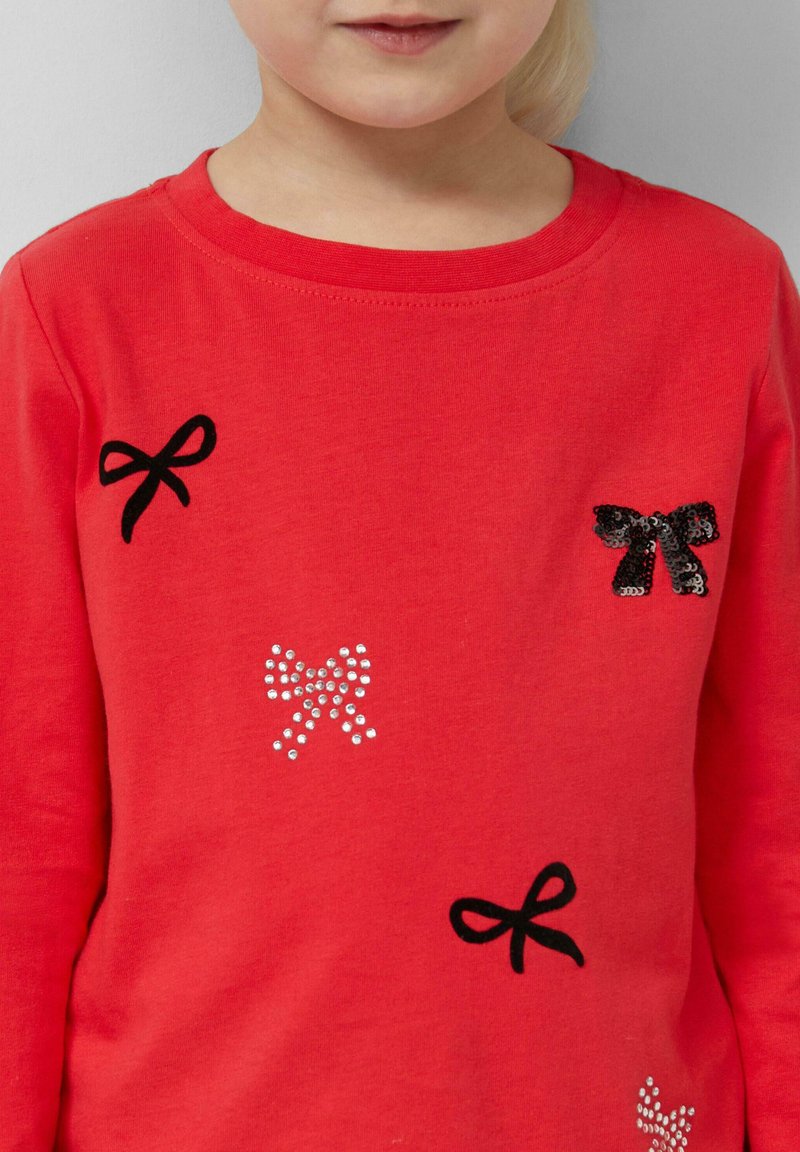 Rotes Baumwollhemd mit langen Ärmeln, das auf der Vorderseite mit schwarz gestickten Schleifen und Kristallakzenten in verschiedenen Größen verziert ist. Glatte Textur, runder Ausschnitt.