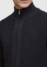 Stickad zip-tröja i mörkgrått med ett texturerat mönster, hög krage och slät dragkedja. Avslappnad passform med ribbad detalj vid kragen.