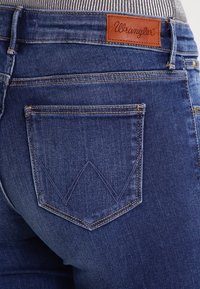 Vybledlé modré džínsy s hnědým koženým označením Wrangler na pase a charakteristickým V-šitím na zadní kapse.