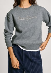 Sudadera gris con puños y dobladillo acanalados, adornada con un logo bordado en blanco. Combinada con pantalones de rayas azul marino.