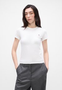 Kvinde med langt mørkt hår iført en tætsiddende hvid kortærmet t-shirt og grå stribede bukser med hænderne i lommerne mod en ensfarvet baggrund.