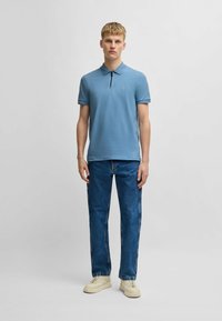 Ljusblå bomullspikétröja med dragkedja i kragen, i kombination med mörkblå jeans med sidostygn och krämfärgade sneakers.