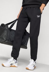 Calças de treino pretas com um cós elástico e o logótipo da Reebok. Combinadas com ténis brancos de desporto com detalhes pretos e solas de borracha.