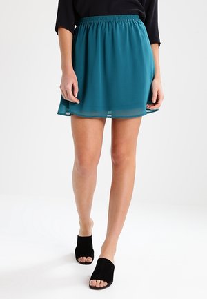 Persoon die een korte teal rok, een zwart topje en zwarte open schoenen met hak draagt, staand tegen een effen witte achtergrond.