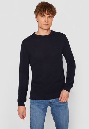 Navy blue knit sweater with a crew neck, long sleeves, textured pattern, and a small white logo on the chest. Worn with light blue jeans.