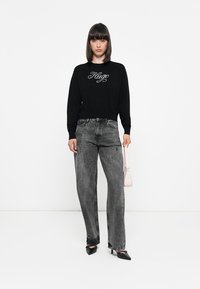 Sudadera negra con "Hugo" bordado en blanco, combinada con jeans de mezclilla gris de tiro alto. Los accesorios incluyen un pequeño bolso rosa y tacones negros.