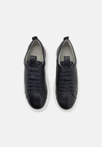 Svarte lær-sneakers med teksturerte tåhatter, flate snørebånd og løftestropper; hvite gummisåler, minimalistisk design med glatte detaljer.