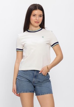 Vrouw draagt een wit shirt met korte mouwen met groene en marineblauwe rand en spijkershorts, staat met één hand in de zak.