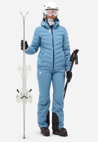 Veste de ski matelassée bleu clair avec des accents noirs, pantalon bleu assorti, gants noirs, casque blanc et équipement de ski, y compris des skis et des bâtons.