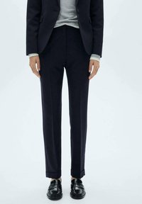 Námořnické kalhoty s upraveným střihem, s hladkou texturou, plochým předním dílem a mírně ohrnutými manžetami. Nosič má na sobě černé boty a šedý top.