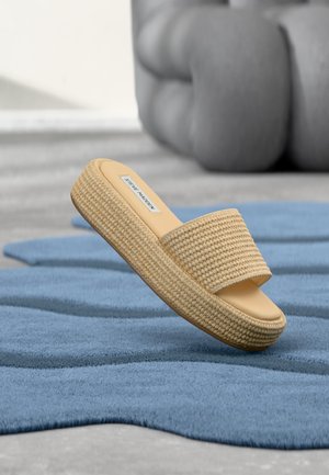 Beige geweven platform slidesandaal met een brede band en Steve Madden-label, zwevend boven een gestructureerd blauw vloerkleed op een grijze vloer.